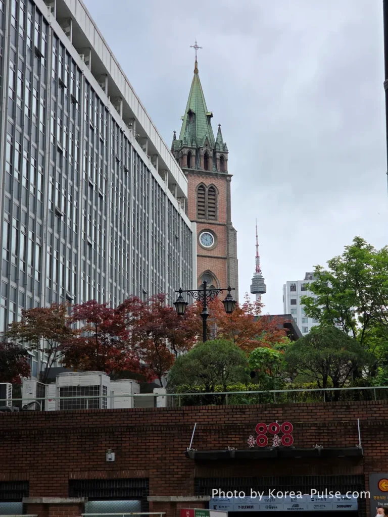 Myeongdong Cathedral Gothic spires rising above modern Seoul buildings near Olive Young Myeongdong Town on a cloudy spring day
