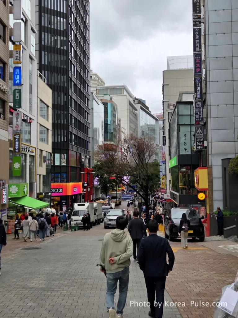 Myeongdong main street sloping downhill with shoppers and tourists, leading to the corner where Olive Young Myeongdong Town flagship is located