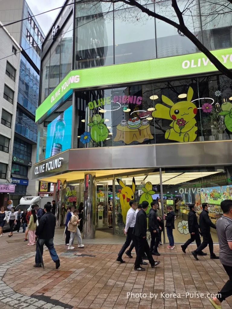 Olive Young Myeongdong Town flagship store full exterior view with Pokémon character collaboration decoration on windows and a crowd of international tourists at the entrance