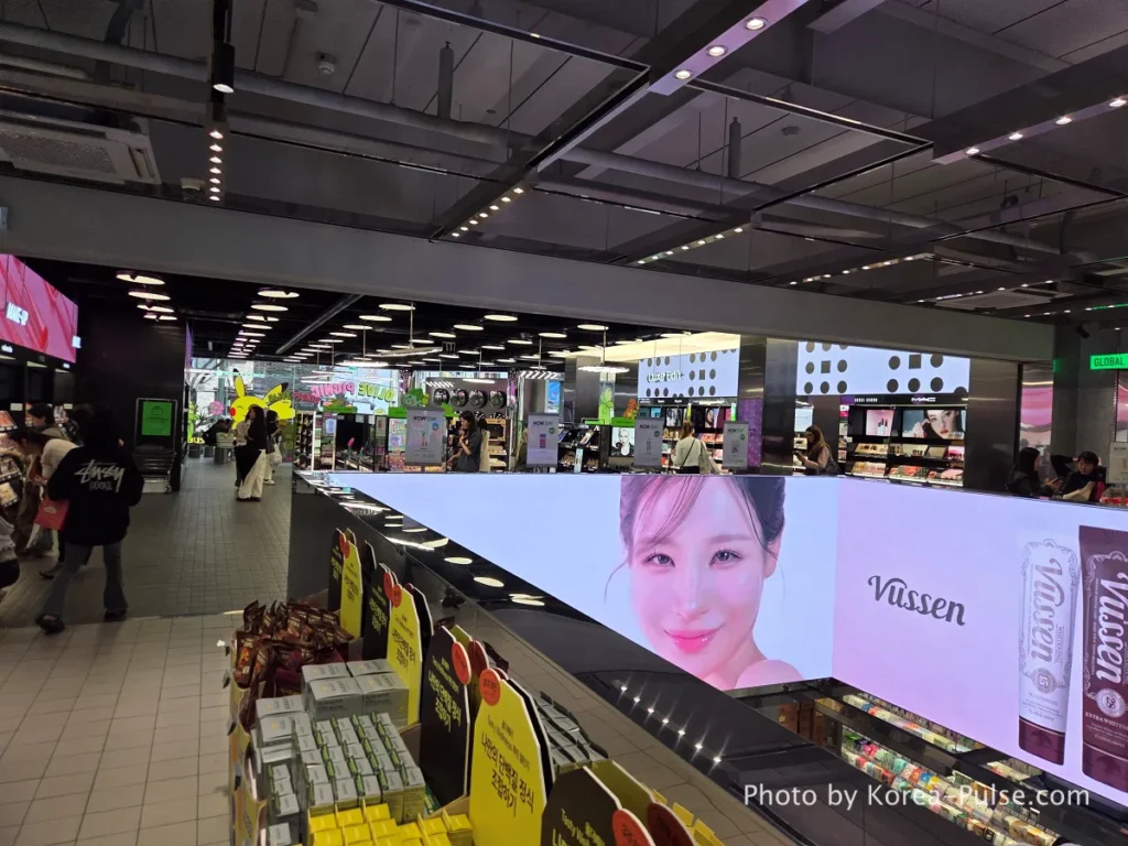 A wide view from the second floor of Olive Young Myeongdong Town, showing a massive digital screen advertising Vussen toothpaste, shoppers browsing makeup sections, and a glimpse of the first floor below.