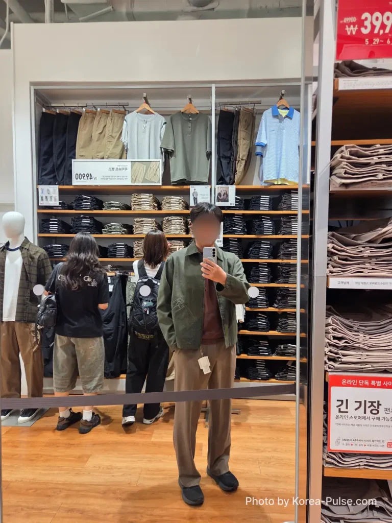 Uniqlo Thank You Festival in Seoul: I Went for One Thing and Bought Another (No Regrets) 8 Mirror selfie of a man trying on the olive green Uniqlo JW Anderson Zip-up Blouson inside a Uniqlo Seoul store, with other shoppers and clothing racks visible in the reflection
