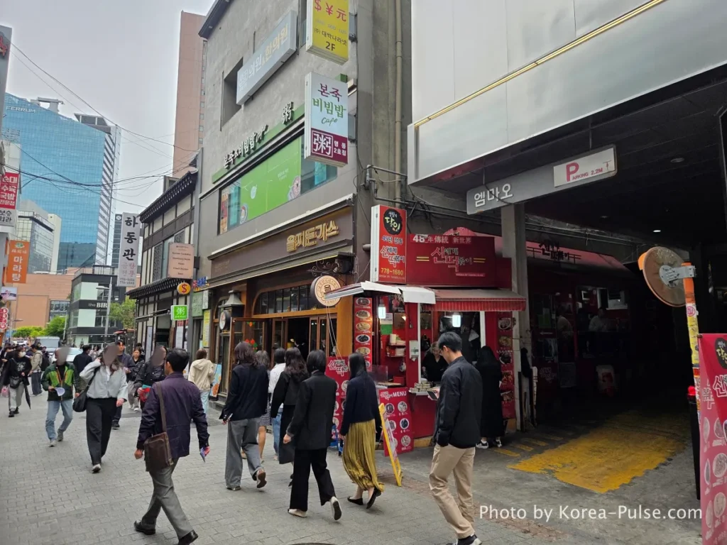 Myeongdong Street in May: Where Seoul's Best Food Hides in Plain Sight 4 Busy Myeongdong street alley at lunchtime with restaurant signs including Eulji Tonkatsu and Hadongkwan visible, pedestrians walking