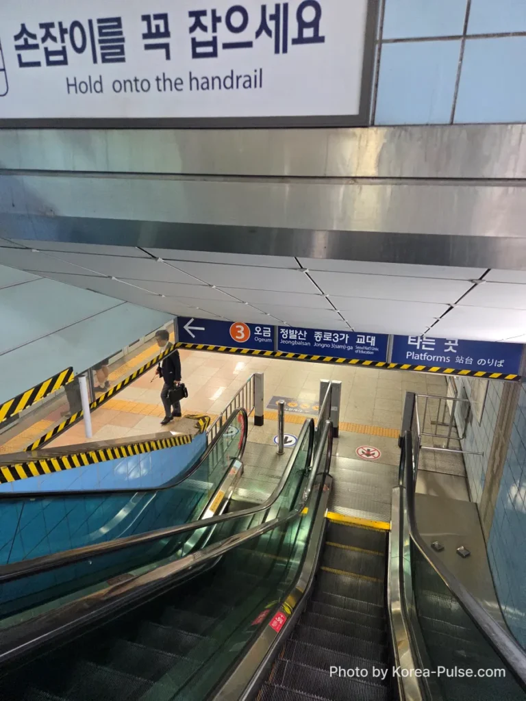 Escalator descending to Seoul Metro platform with bilingual Korean and English safety sign reading "Hold onto the handrail"