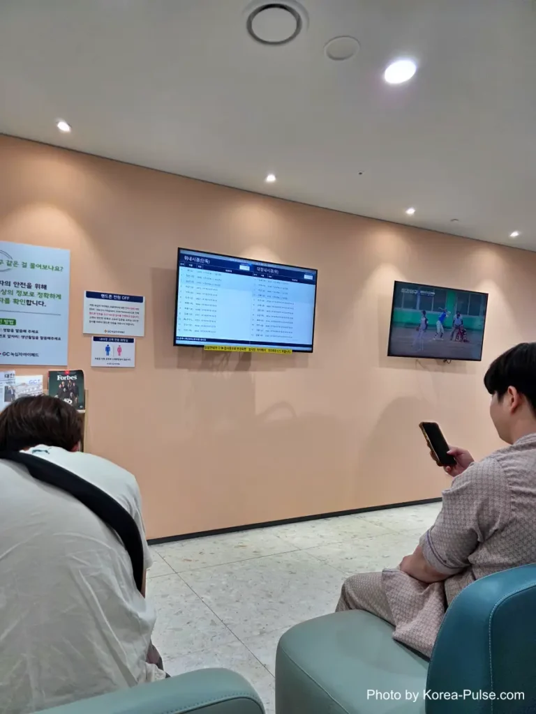 Inside Korea's 2-Hour Health Checkup Machine: Wristbands, Yellow Lines, and a Bowl of Porridge 12 Crowded waiting room with a digital queue monitor listing patient names for the endoscopy department